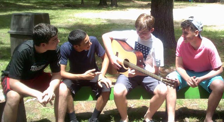 teenage boy camper playing guitar on bench with friends